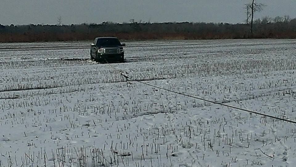 Black truck in snowy field, pulling line, trees in background.