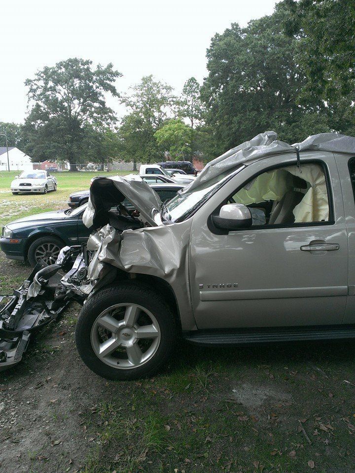 Damaged silver SUV with deployed airbags, debris scattered. Parked on dirt near other vehicles and trees.