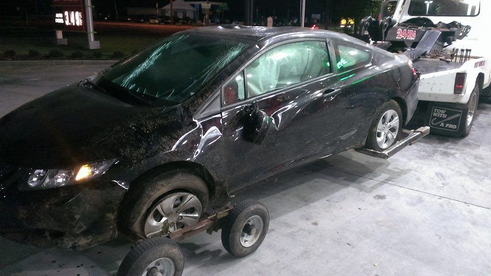 Black car, damaged, being towed. At gas station, daytime.