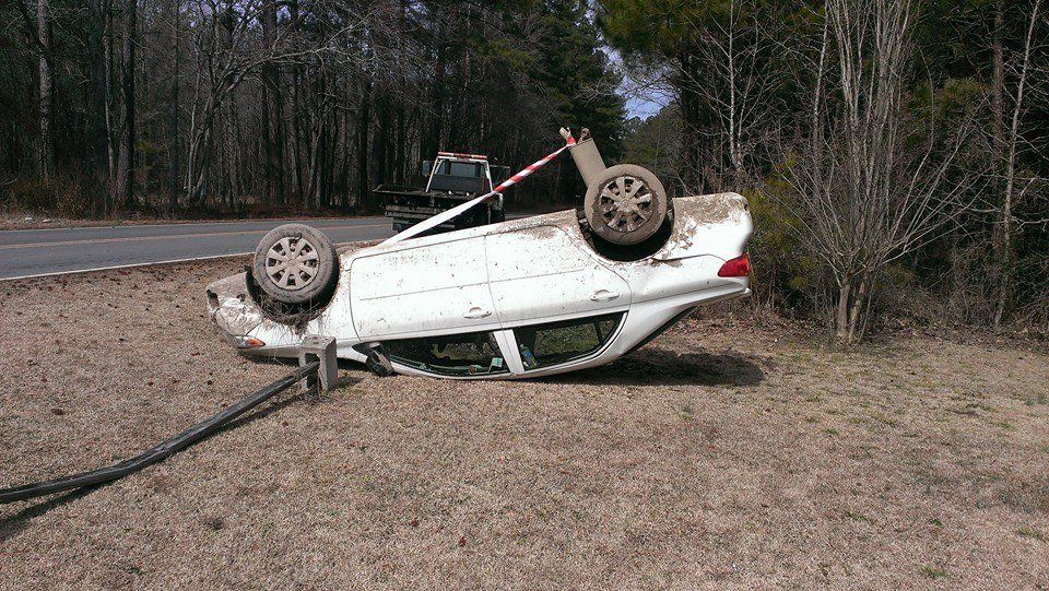 White car overturned on side of the road in a grassy area, possibly after a crash.