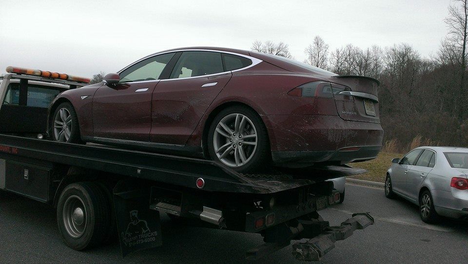 Maroon Tesla sedan being towed on a flatbed truck; grey car in the right lane.