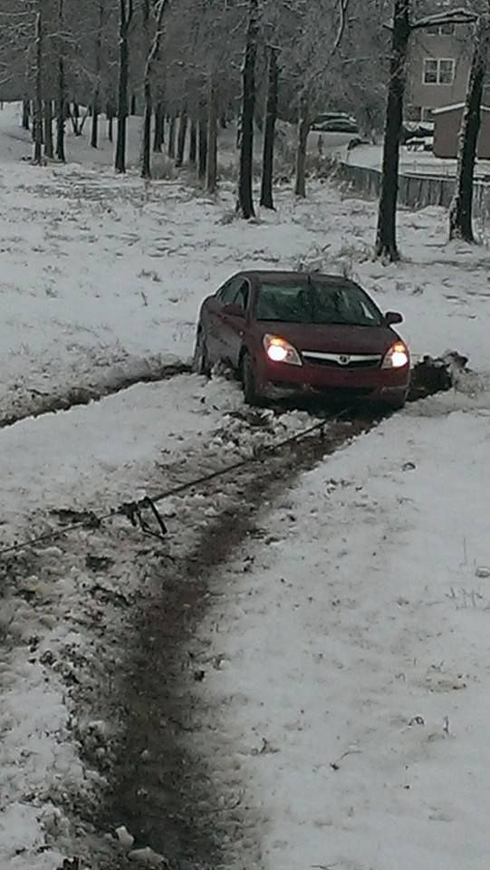 Red car stuck in snow-covered dirt tracks, in a snowy field beside trees.