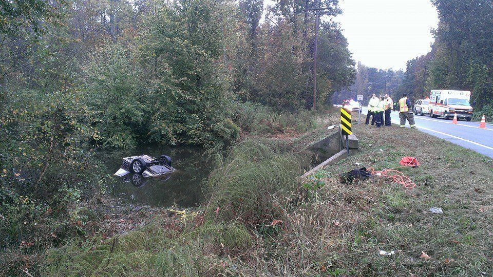 Car submerged in water next to a road, emergency personnel standing nearby.