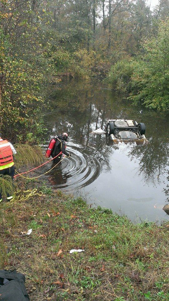 Car submerged in water, rescue diver in water, another person on bank. Overcast, wooded background.