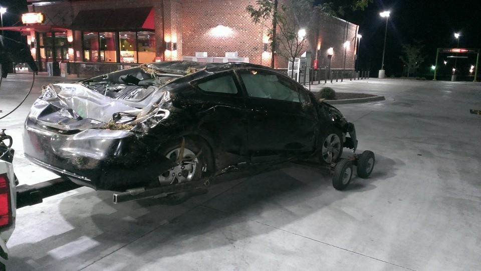 Black car heavily damaged and towed at a gas station at night.