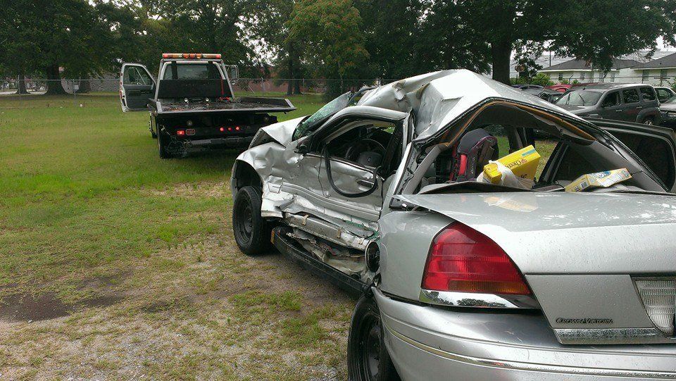 Damaged silver car, roof crushed, with tow truck on grassy area, indicating accident.