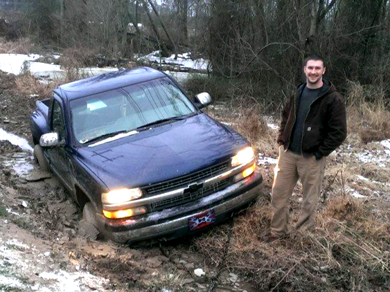 Man smiles beside a blue pickup truck stuck in mud and snow.