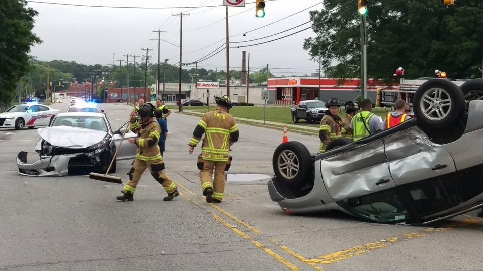 Firefighters at a car accident scene with a flipped vehicle and damaged police car on a road.