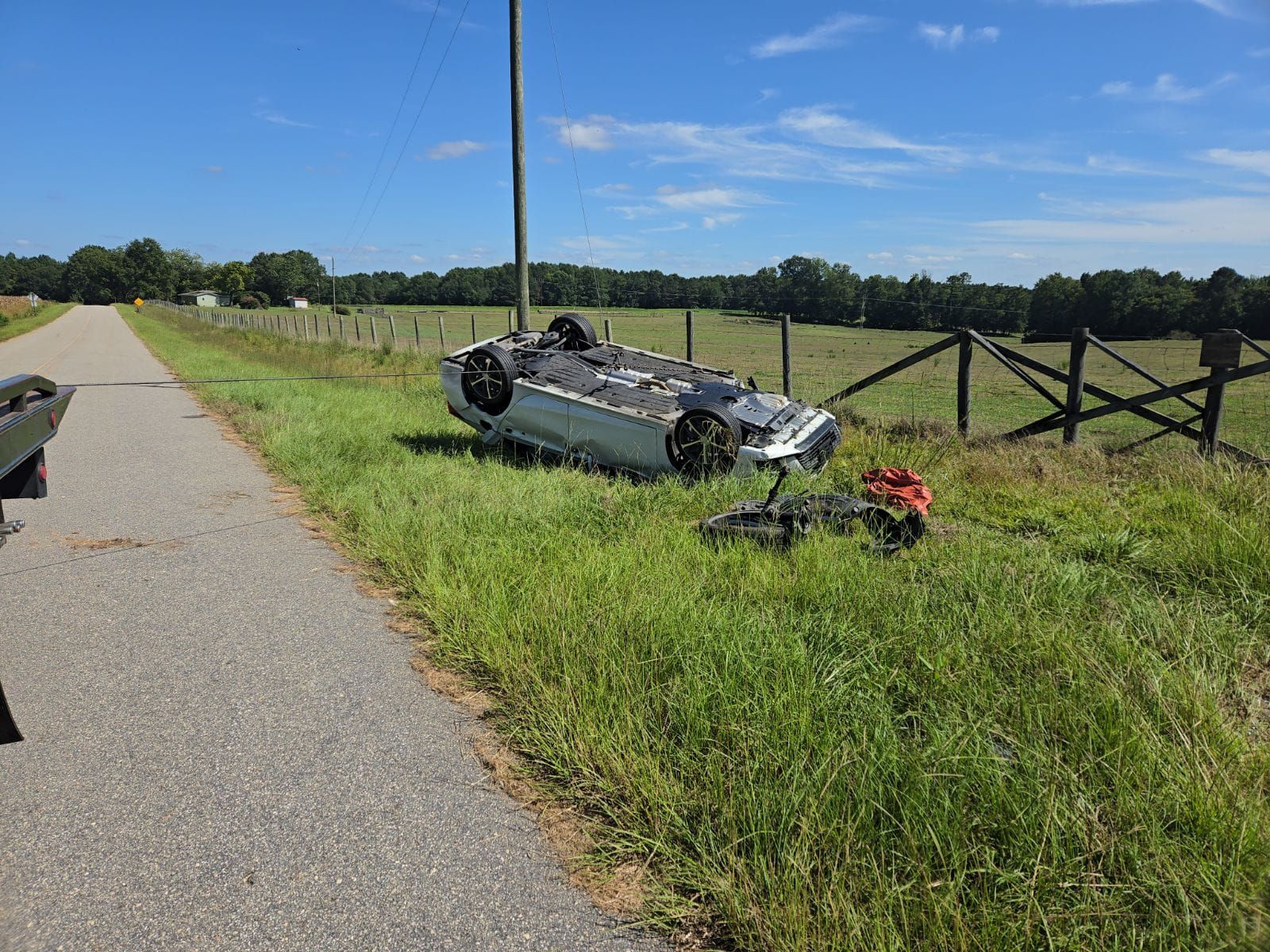 Car overturned in grassy ditch next to road, fence and telephone pole in background, sunny day.