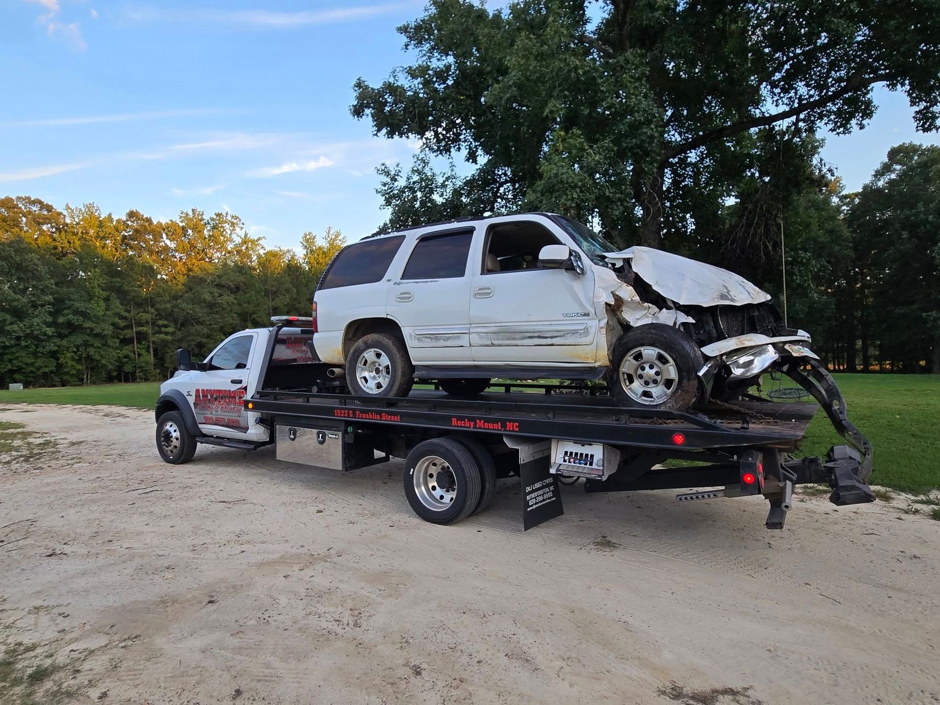 White SUV, heavily damaged in a collision, being towed on a flatbed truck on a dirt road.