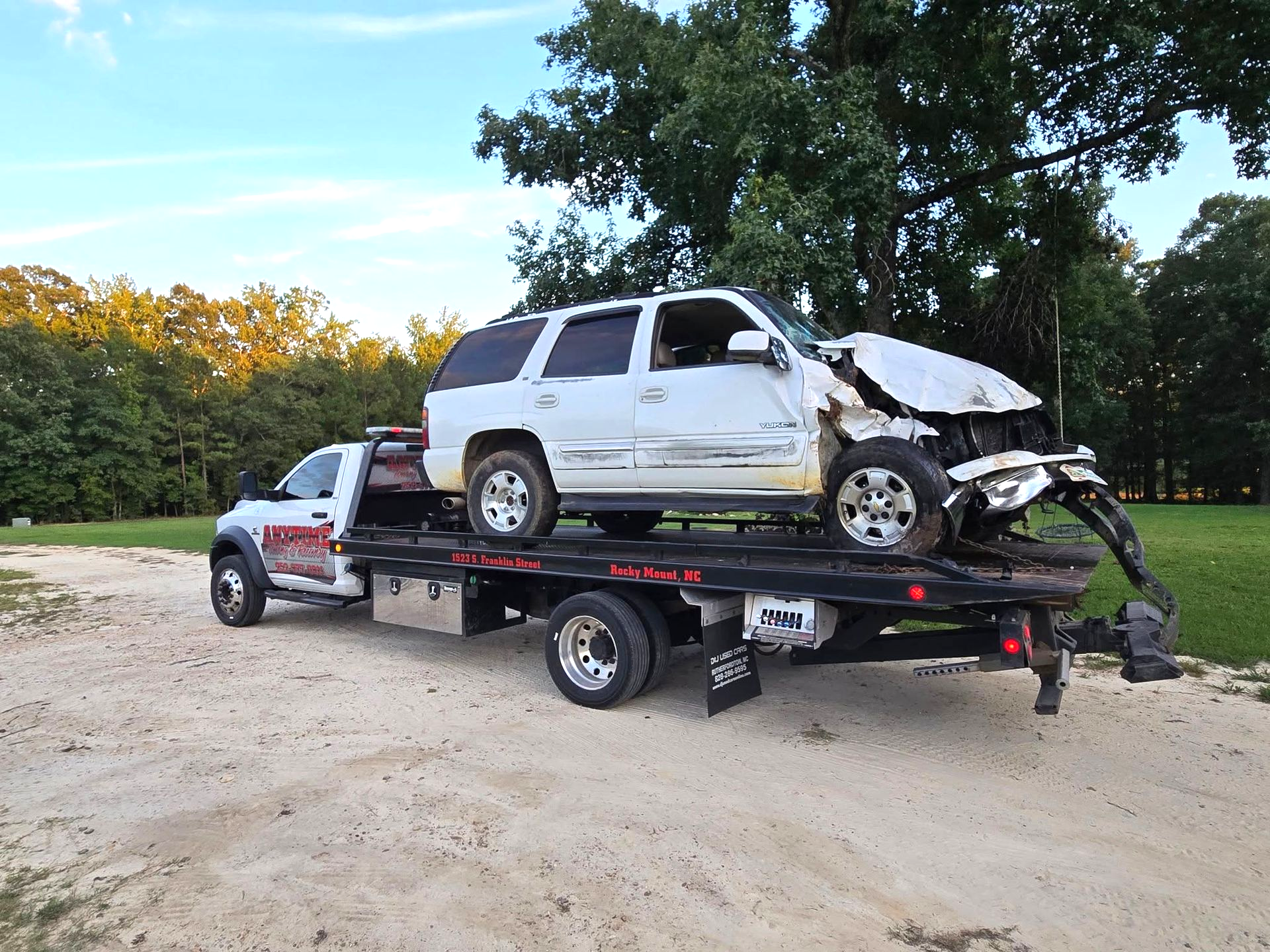 White SUV damaged in accident, loaded onto a flatbed tow truck on a dirt road.
