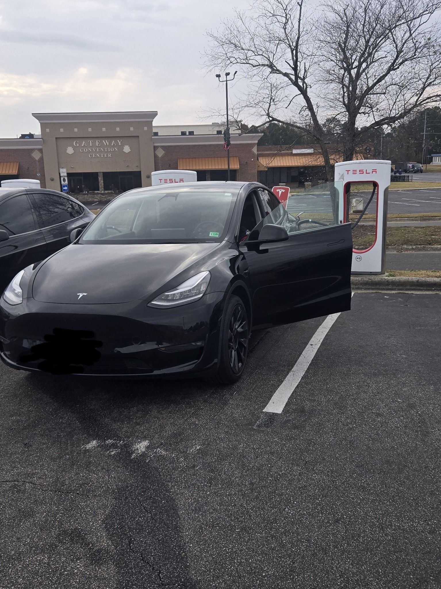Black Tesla Model Y parked near a charging station, door open, in front of a shopping center.