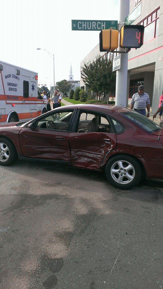 Damaged maroon car at an intersection with an ambulance; people nearby. 