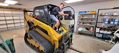 A man in a black shirt and blue shorts is inside a yellow skid steer in a workshop.