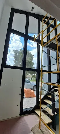 Interior view with a tall window, stairs, and a scaffolding. The window shows blue sky and trees.