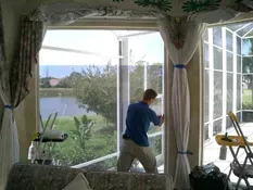 Man installing window screen overlooking lake, curtains draped on either side.