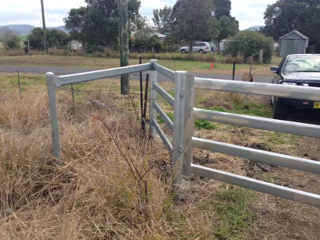 A corner section of a galvanized steel post-and-rail farm fence set against a rural grassy background.