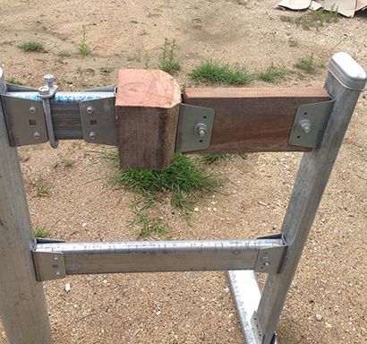 A metal road safety barrier segment featuring a wooden block spacer, mounted on two vertical posts on a dirt surface.
