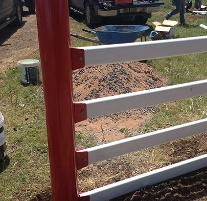 A red metal fence post with white horizontal rails installed in an outdoor area, with a gravel pile and truck in back.