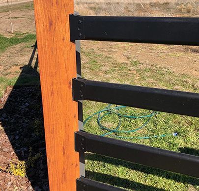A close-up view of a wooden fence post with four black horizontal rails attached by metal brackets in an outdoor setting.