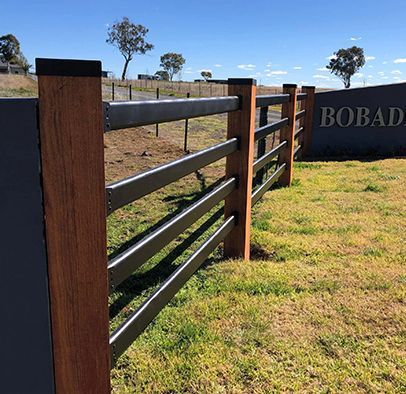 A modern fence with wooden posts and black horizontal rails next to a stone sign in a grassy field under a blue sky.