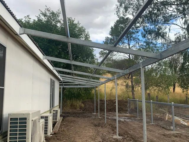 A metal pergola framework under construction attached to the side of a white building with outdoor air conditioning units.
