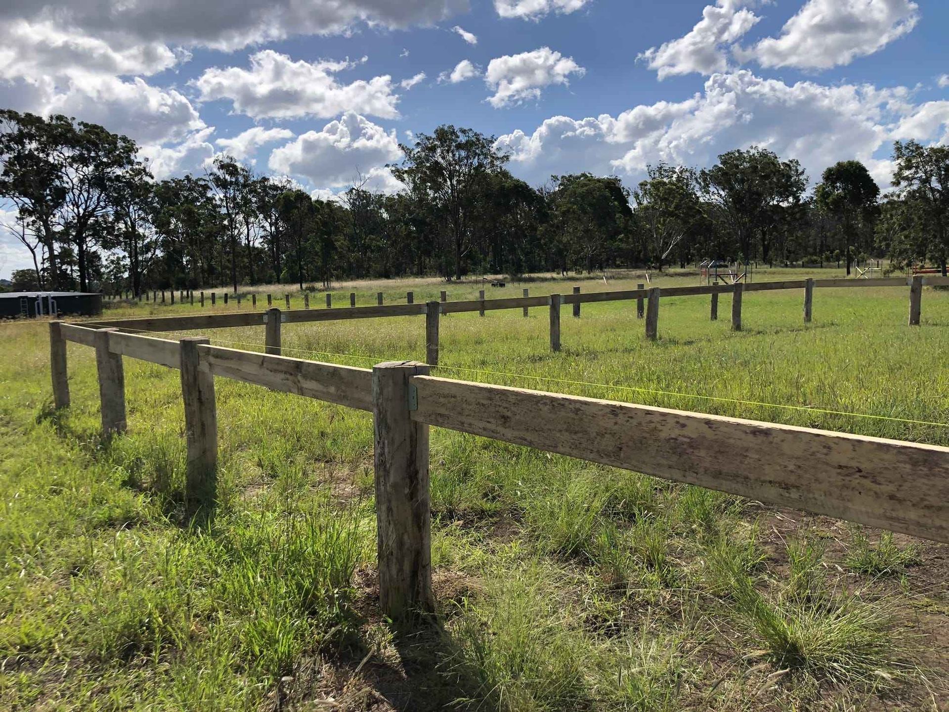 A wooden post-and-rail fence encloses a grassy field with trees under a blue sky with scattered clouds.