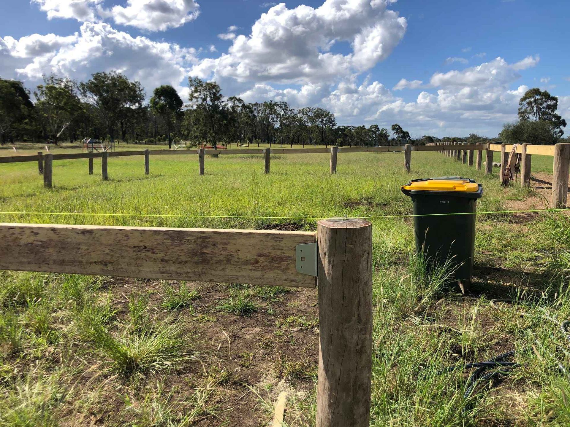 A grassy field surrounded by a wooden post-and-rail fence under a partly cloudy blue sky, with a yellow-lidded bin nearby.
