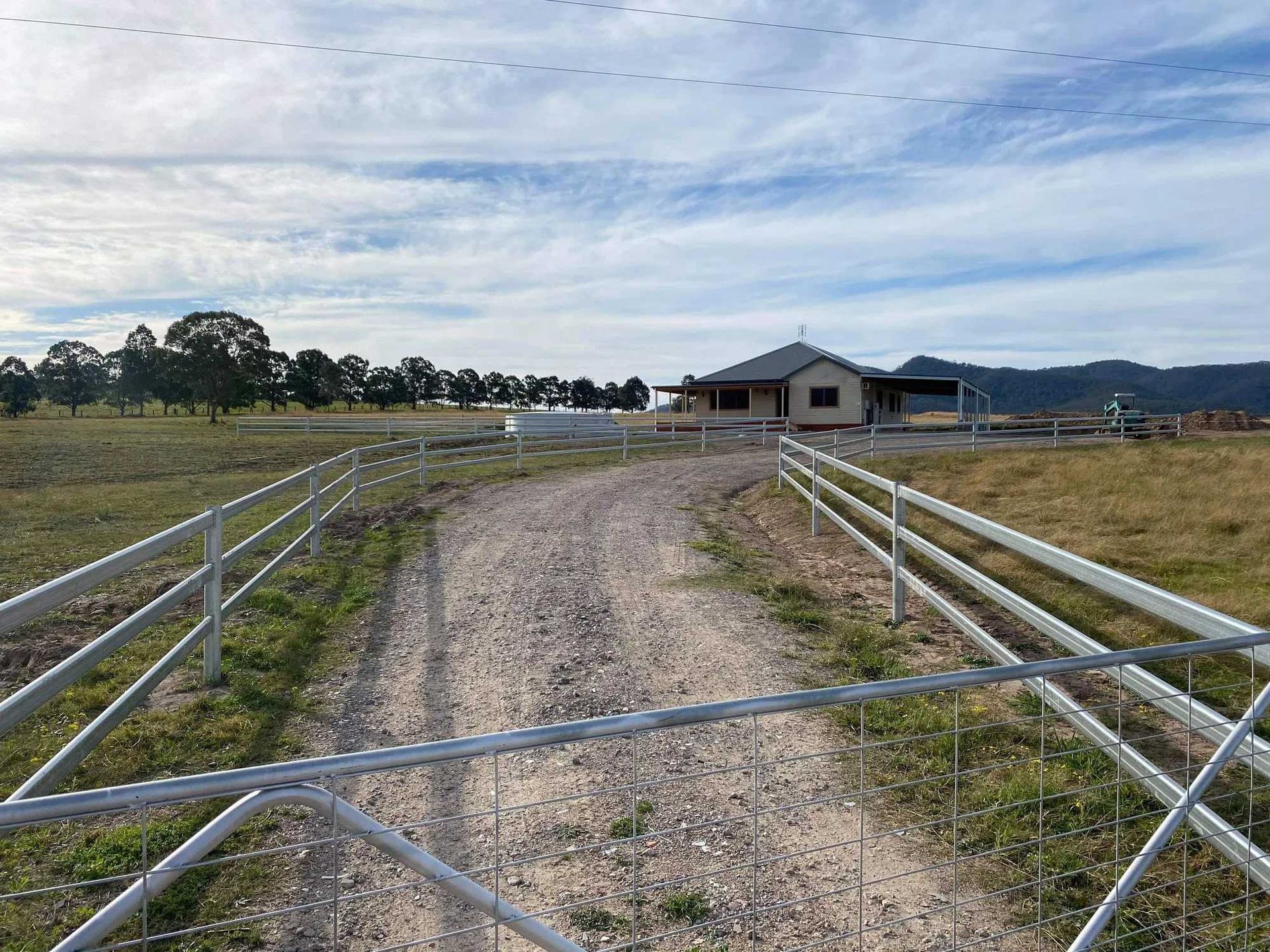 A gravel driveway leads through a metal gate toward a single-story house in a rural, grassy field under a cloudy sky.
