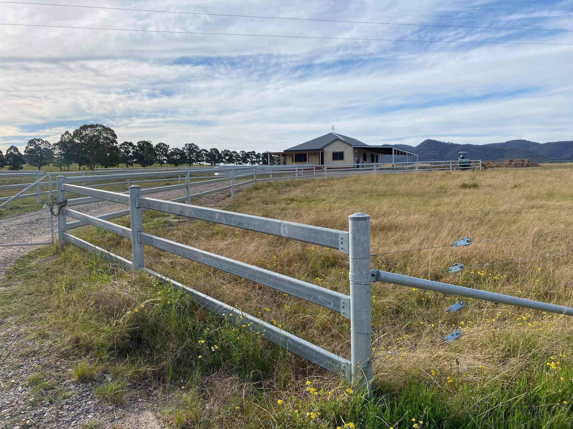 A metal fence borders a dry, grassy paddock leading to a single-story house under a cloudy sky.