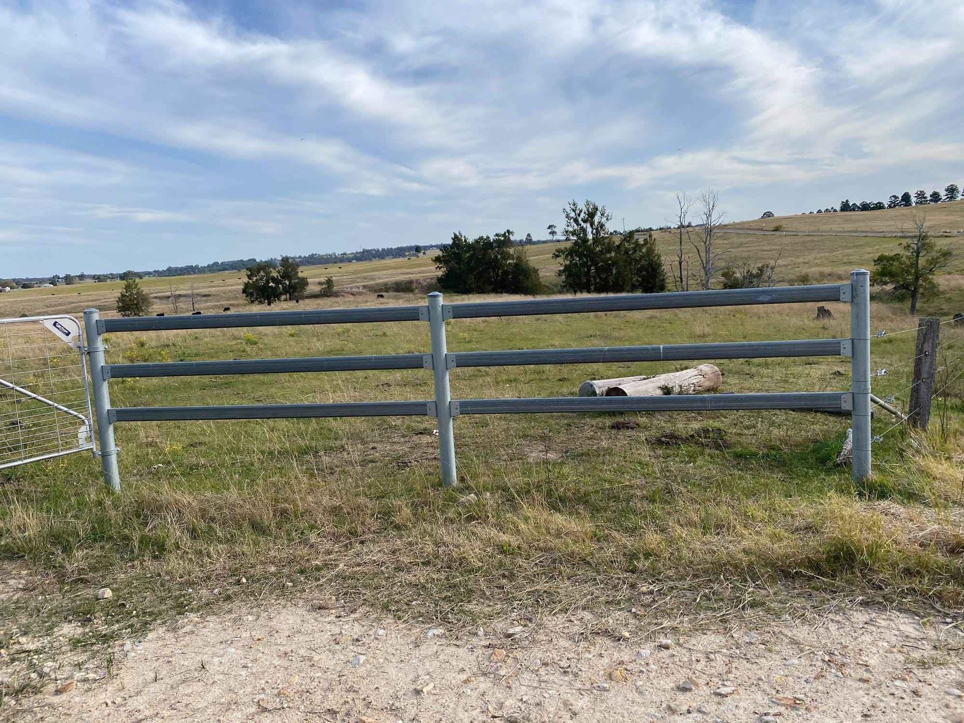 A three-rail metal fence stands in a grassy field under a bright, cloudy sky.