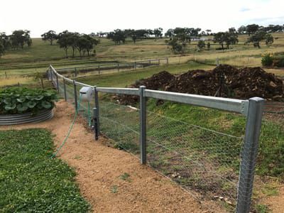 A metal fence with wire mesh runs through a rural yard with a pile of dirt and distant fields in the background.