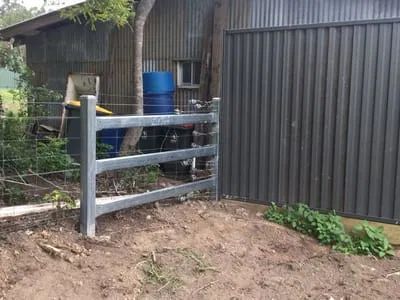 A grey steel fence post with horizontal rails sits on a dirt patch near a corrugated metal wall and wire mesh fencing.