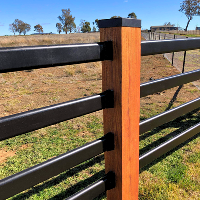 A wooden fence post with a black cap supports four horizontal black rails in a sunny, rural pasture.
