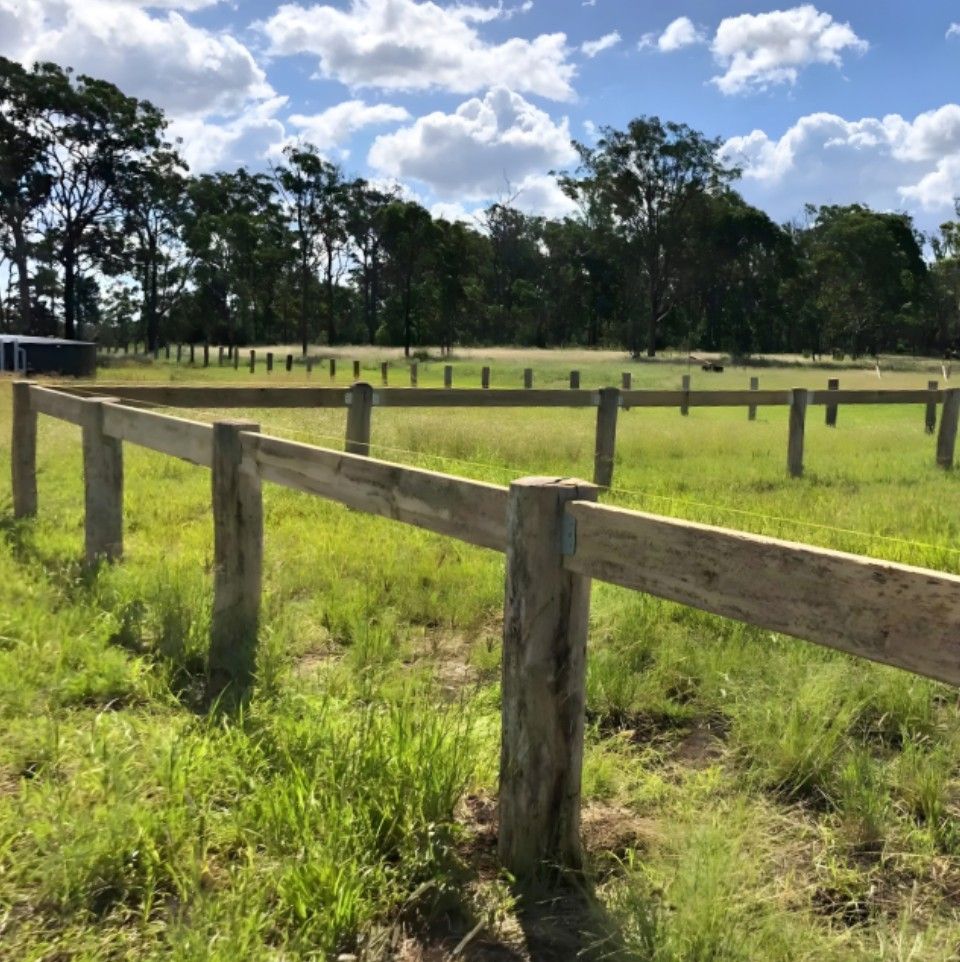 A wooden post-and-rail fence encloses a grassy field under a sunny, partly cloudy blue sky with trees in the background.