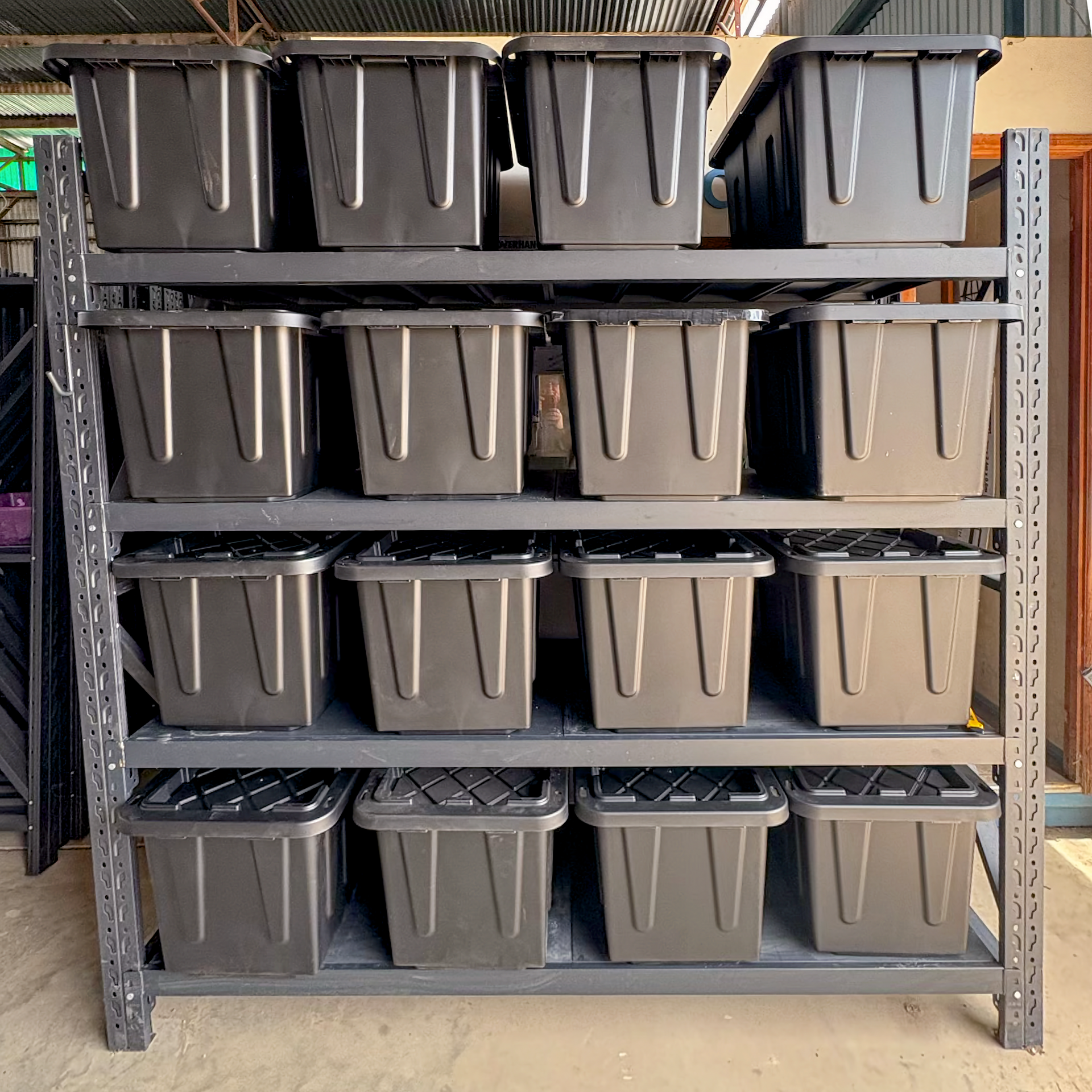 A metal shelving unit with four levels, each containing four identical black storage bins with lids.