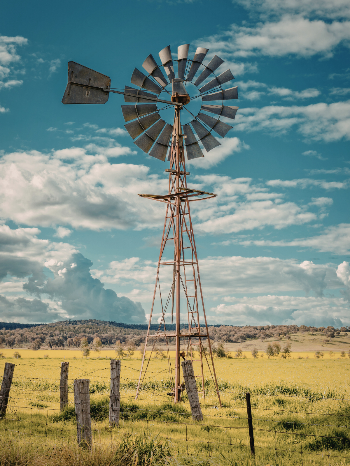 A weathered windmill stands in a vast, dry, golden field under a blue sky with white, scattered clouds.