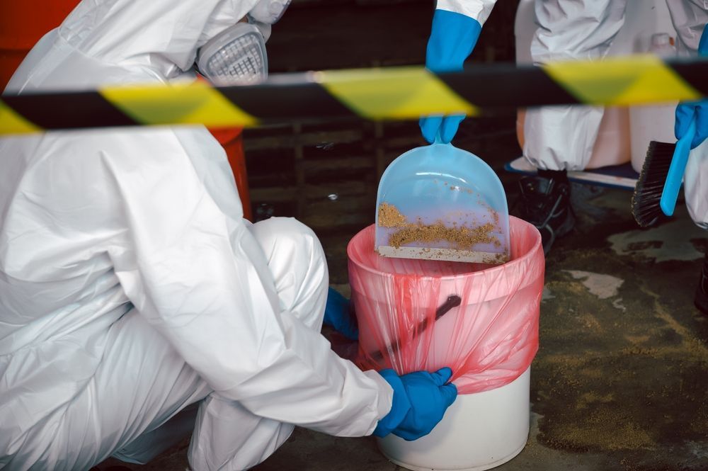 A person in a protective suit is cleaning a trash can with a shovel.