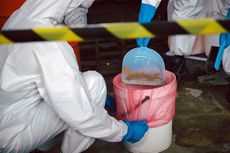 A person in a protective suit is cleaning a trash can with a shovel.