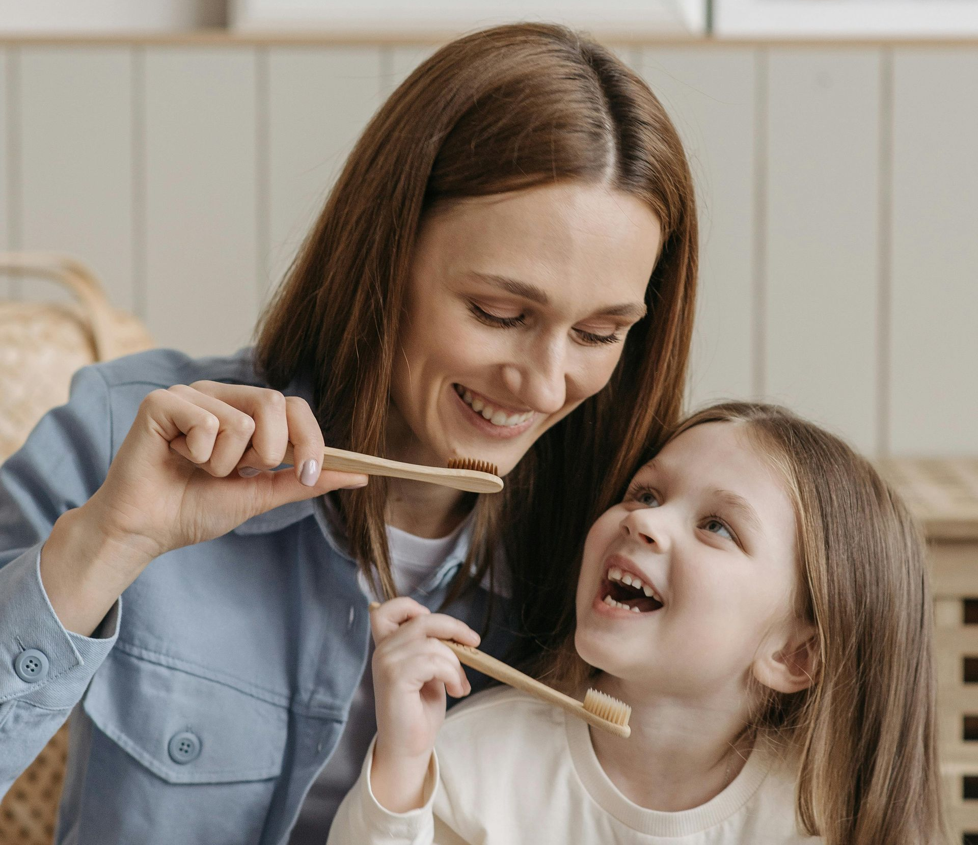 A woman smiling as she shows a child how to brush teeth with wooden toothbrushes. Indoor setting.