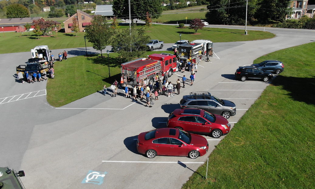 Firetruck surrounded by people in a parking lot. Cars parked nearby. Green grass and trees in the background.