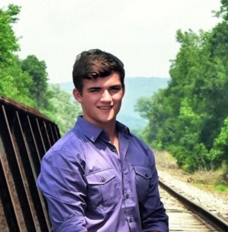 A young man in a purple shirt is standing on train tracks