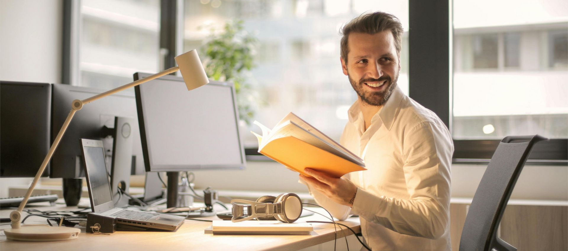Een glimlachende man met een open boek in zijn handen zit aan een bureau met computer, lamp en koptelefoon.