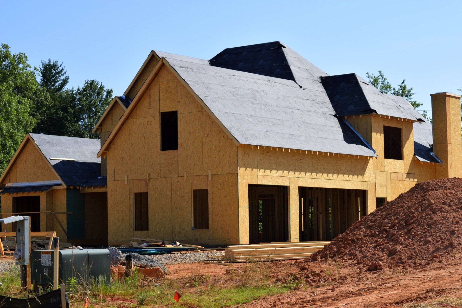 A partially constructed two-story house with wooden framing, dark roofing underlayment, and a dirt pile in the foreground.