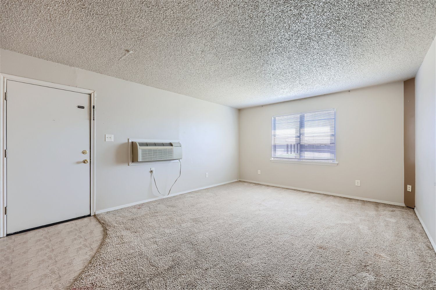 Empty apartment room with carpet, closed door, window with blinds, and air conditioner.