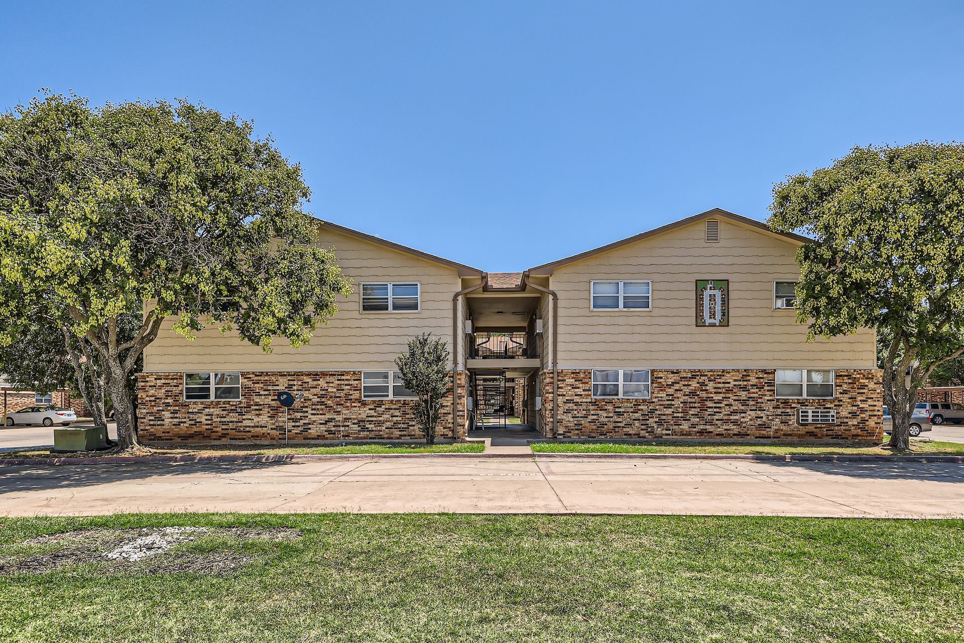 Two-story tan and brick apartment building with green trees against a clear blue sky.