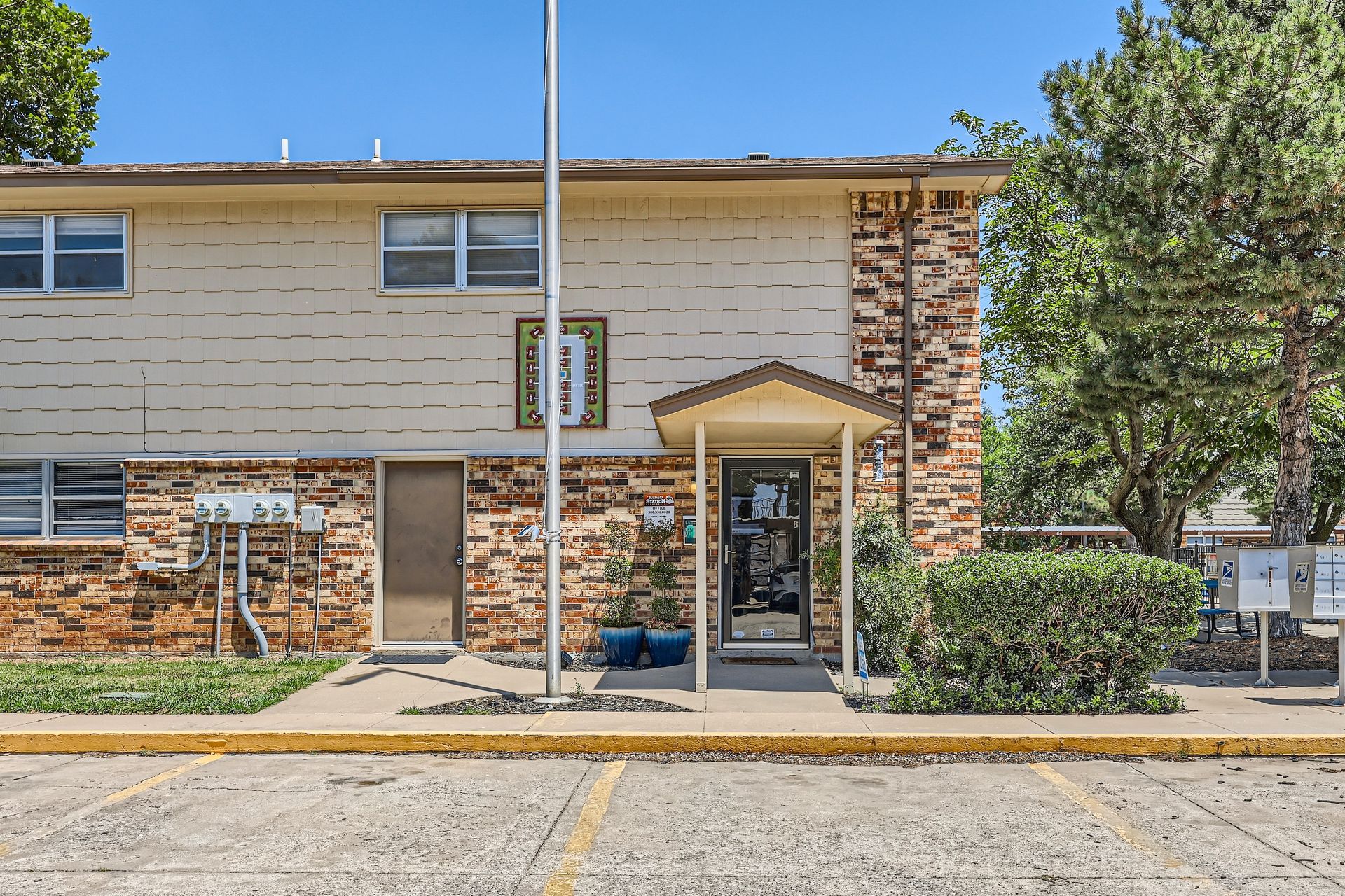 Two-story tan apartment building with brick accents and a small porch entrance under a clear blue sky.