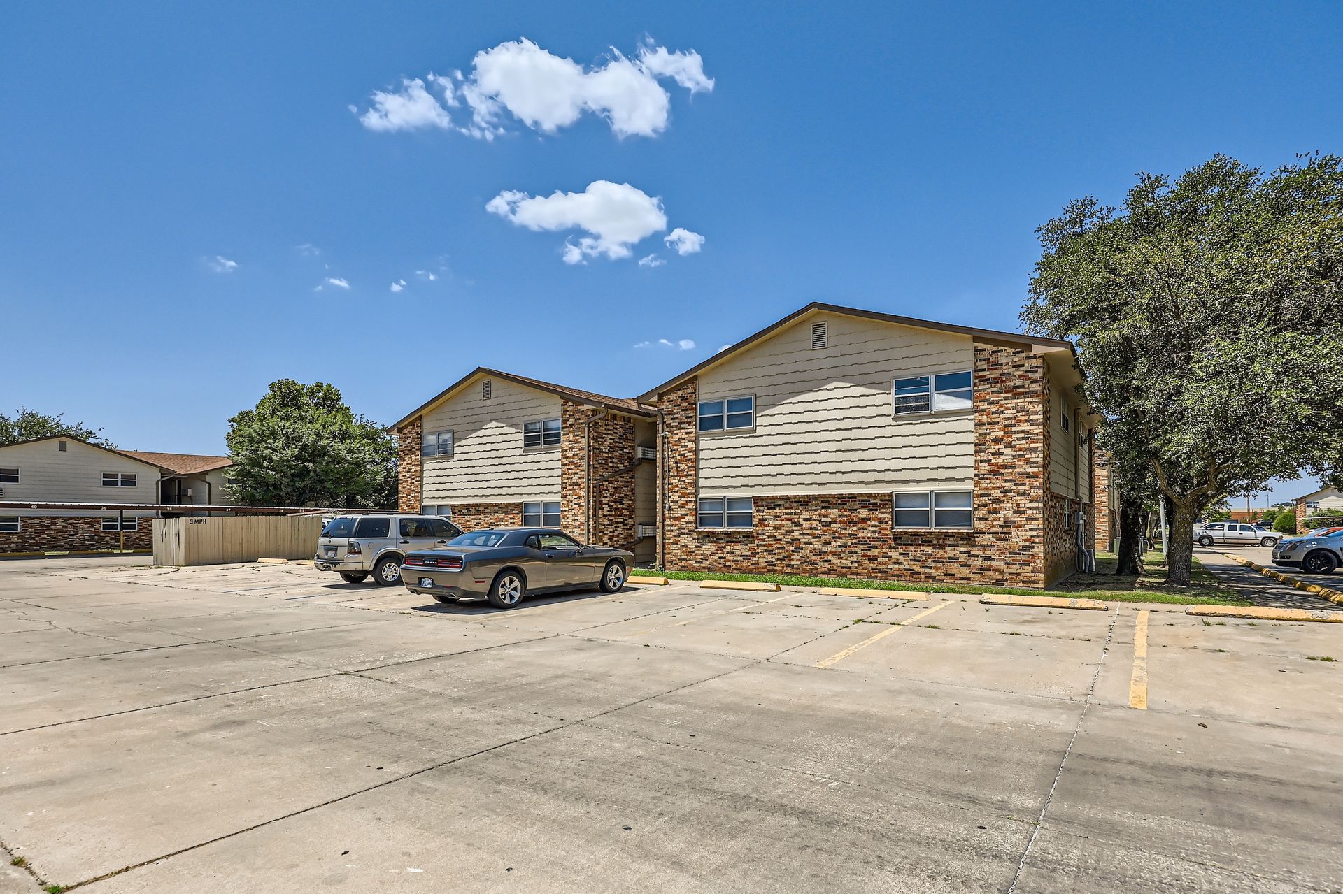 Two-story brick apartment buildings with cars parked in front on a sunny day.