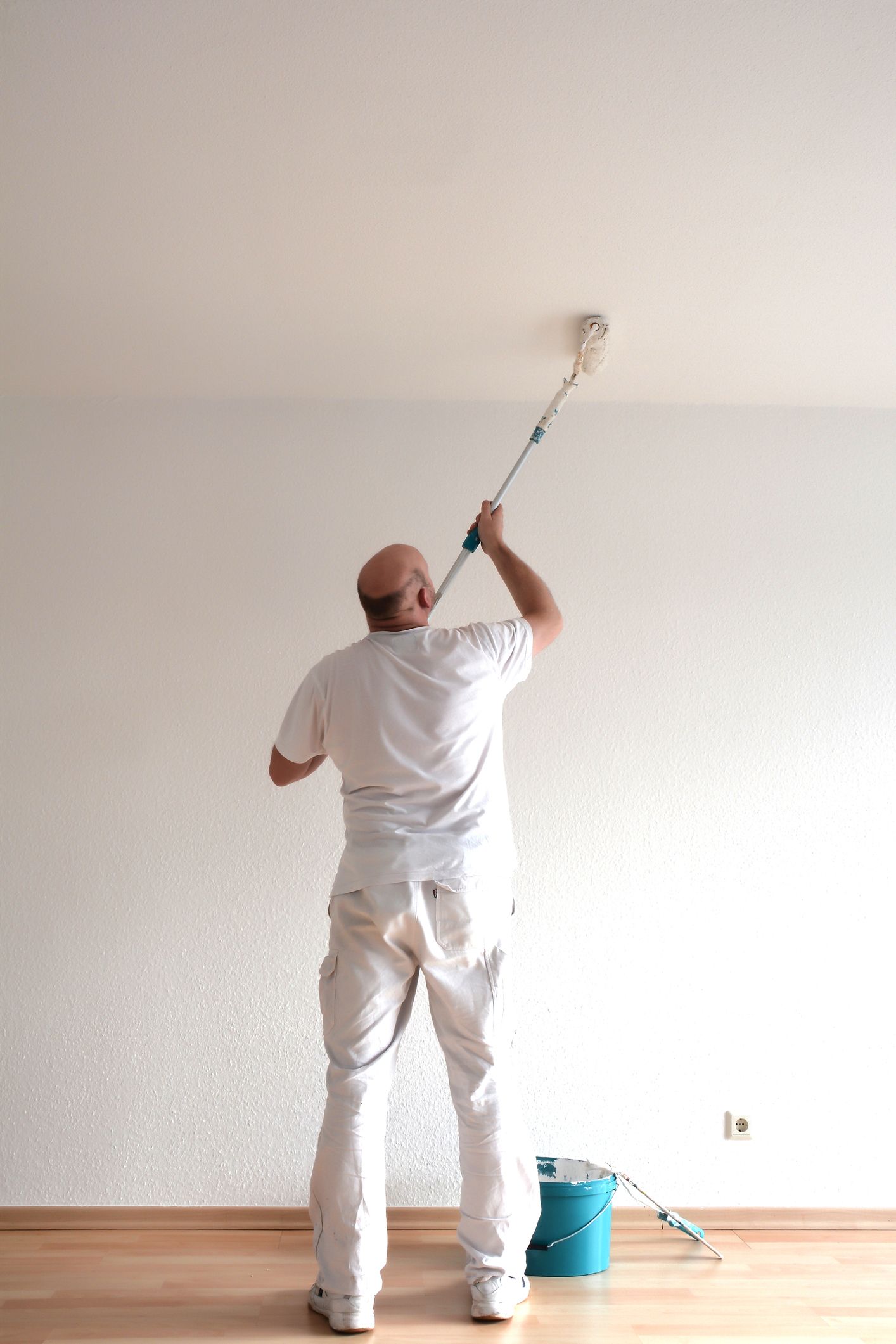 Male painter wearing white clothes paints a room’s ceiling with a paint roller.