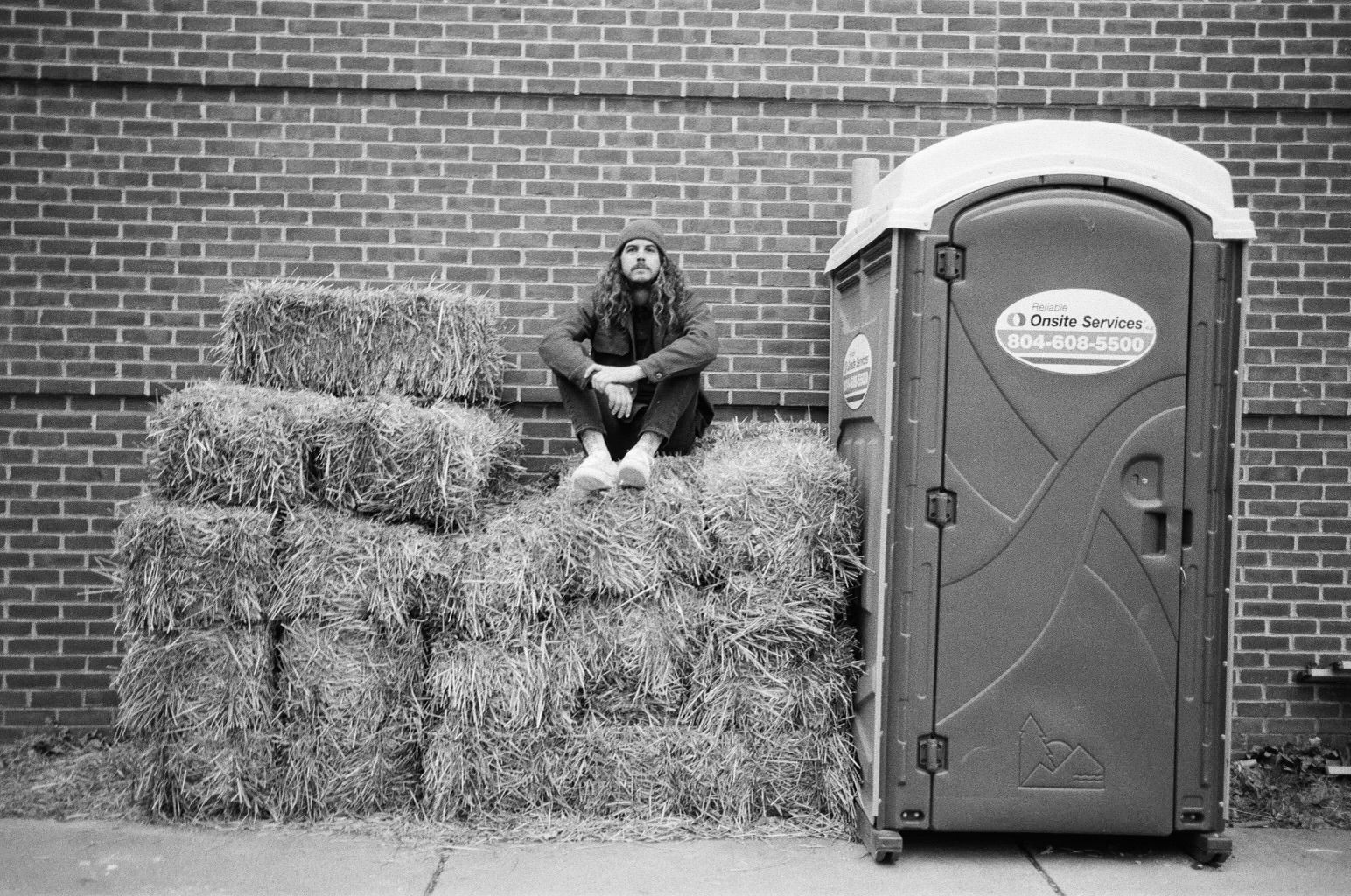 A man is sitting on a pile of hay next to a portable toilet.